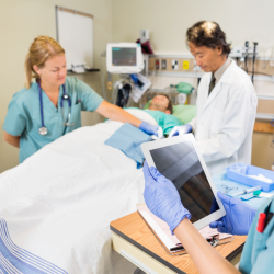 Pasient ligger i sykehusseng med lege og sykepleier ved sin side. Sykepleier holder nettbrett.